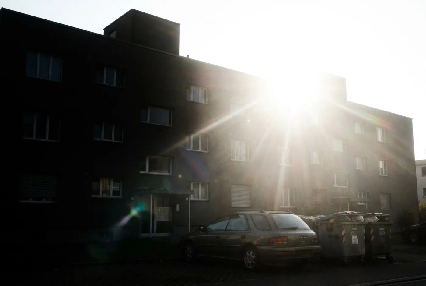 Mehrfamilienhaus gegen die Sonne fotografiert, mit Auto und Mülltonnen vor dem Eingang.