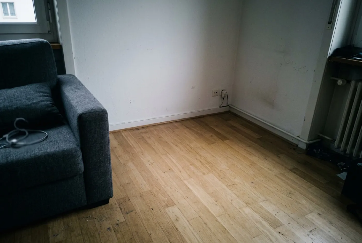 Wohnzimmer aus der Raummitte gegen eine leere Wand fotografiert, dunkel und eng wirkend.